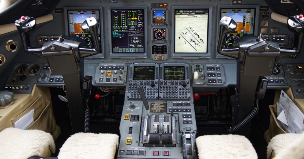 Cockpit with various control panels, buttons, and screens, featuring two pilot seats covered in white fabric.