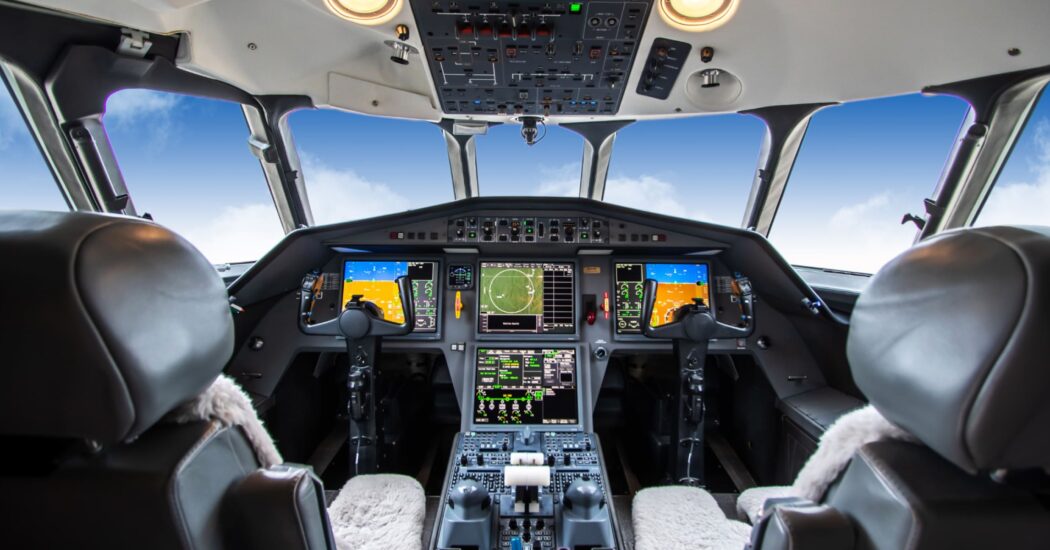 View inside an airplane cockpit with multiple control panels, screens, and two pilot seats, against a backdrop of blue sky and clouds.