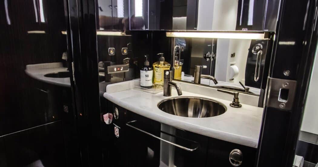 Modern airplane lavatory with a sleek sink, faucet, toiletries, and mirrors. Dark wood paneling and bright lighting.