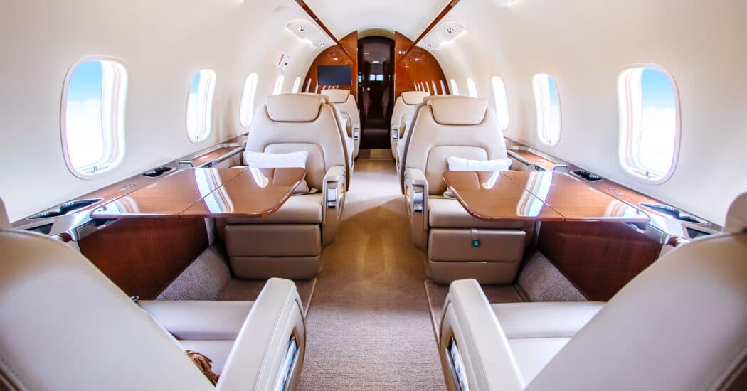 Interior of a luxurious private jet, Bombardier Challenger, with cream leather seats, polished wooden tables, and large windows on both sides.