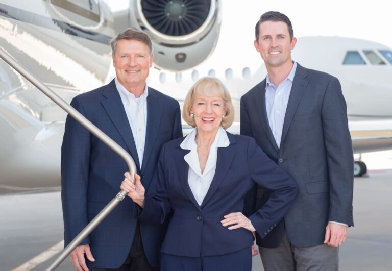 Three people in business attire stand smiling in front of a private jet.