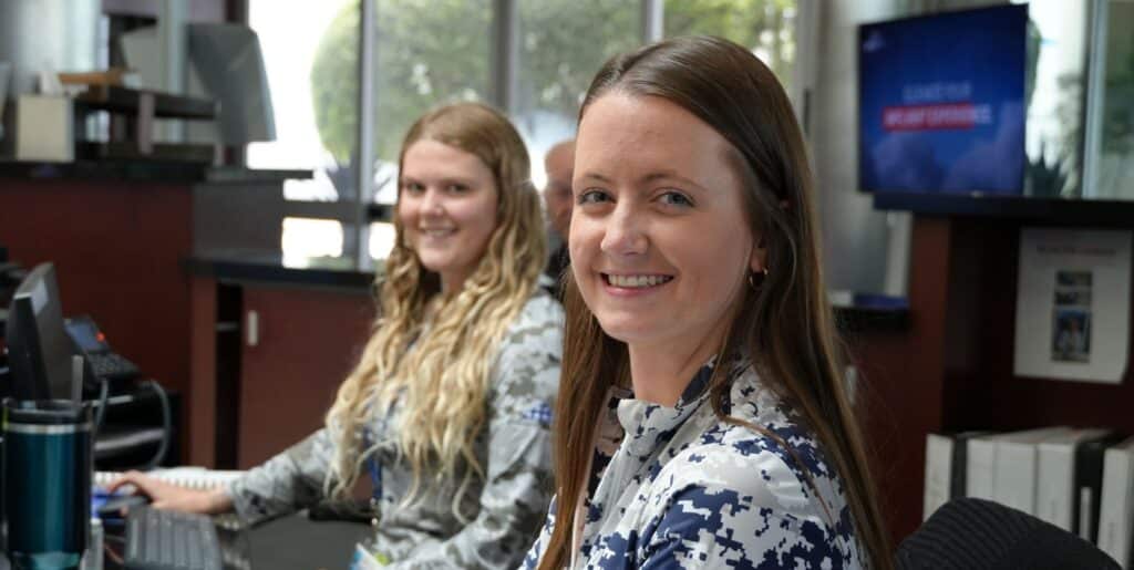Two women sitting at a desk, smiling at the camera, with computers and office materials in the background.