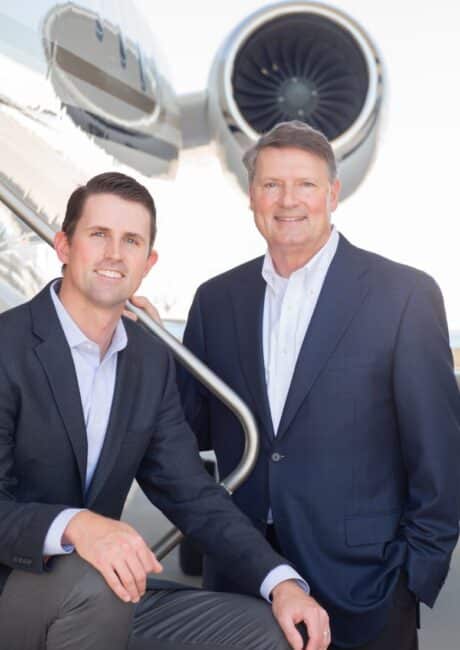 Two men in business attire pose in front of a private jet, one leaning on the stair rail and the other standing beside him.