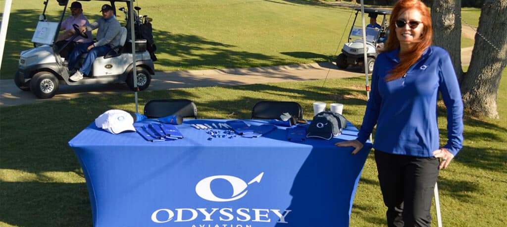 A woman stands in front of a branded table at the Business Jet Cares Charity Golf Tournamant
