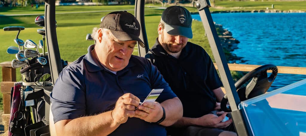 Two golfers sitting in a golf cart recording their score with a scenic golf course and pond behind them