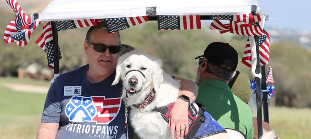 A golf cart decorated with American flags driving away with a Veteran and his Patriot Paws Service dog sitting next to him